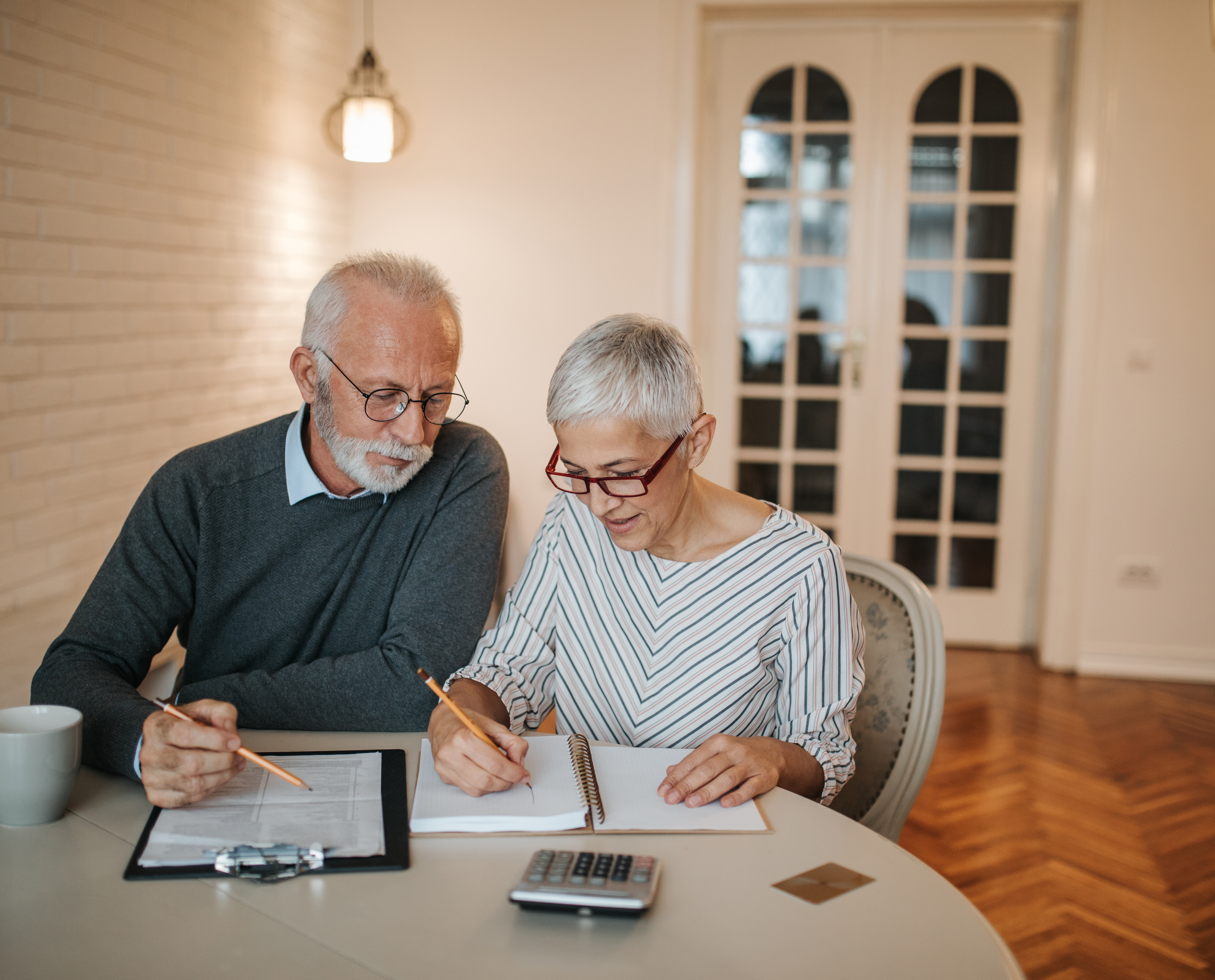 An elderly couple sits at a table in front of a computer and a calculator.