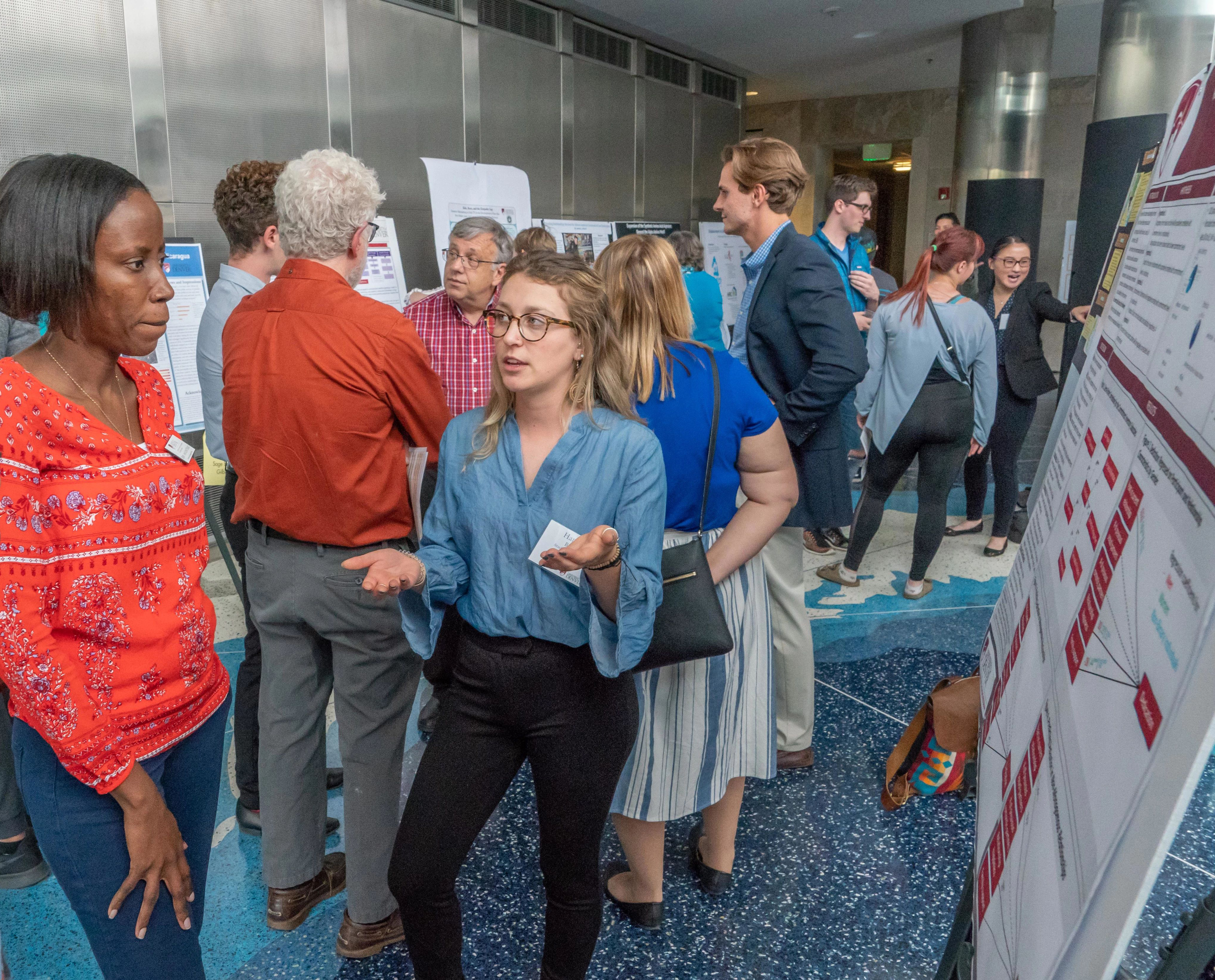 A young woman presents a trifold board research project to several onlookers.